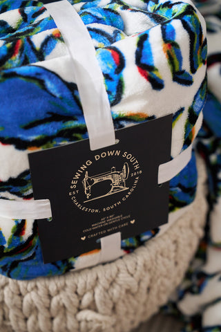 A rolled multicolored fabric blanket features abstract blue, green, and orange patterns. A black label with Sewing Down South and a vintage sewing machine illustration is attached with white ribbon. This Crab Craze, Shades of Blue Blanket embodies coastal chicness against a knitted-texture background.