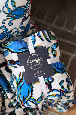The cozy, multicolored Crab Craze, Shades of Blue Blanket showcases a blue and green pattern with a white ribbon. Featuring a Sewing Down South label from Charleston, it’s soft, fluffy, and coastal chic on a brown brick surface.