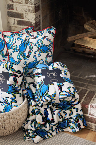 The cozy scene showcases blue and white pillows and a Shades of Blue Blanket from the Crab Craze collection, all tucked in a woven basket by a brick fireplace with stacked firewood, perfecting Sewing Down Souths coastal chic style.