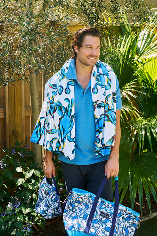 A man in a blue shirt stands outdoors holding two patterned bags, embodying Sewing Down Souths creativity. The Crab Craze Beach Towel, with its vibrant blue and green design, is draped over his shoulders against a backdrop of lush greenery and wooden fencing.