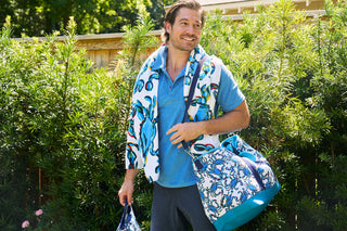 A smiling man with short dark hair stands outdoors in front of lush greenery. Wearing a blue shirt, he drapes a Sewing Down South Crab Craze Beach Towel over his shoulder and holds a patterned item in one hand, along with a matching bag in the other.