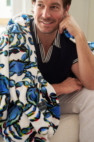 A man smiles while sitting on a couch, wrapped in a Crab Craze, Shades of Blue Blanket. Wearing a navy shirt with a striped collar and white pants, he embodies coastal chicness. The cozy setting enhances the relaxed vibe.