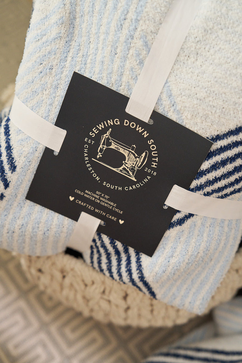 Close-up of the Mod Geo Diamond Luxe Knit Blanket, a striped blue and white plush blanket featuring a black tag with a sewing machine graphic. The tag reads Sewing Down South Modern Collections, including location details and care instructions in elegant typography.
