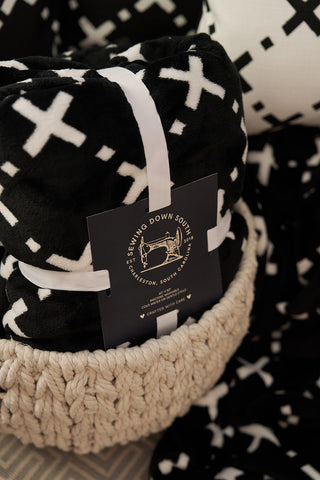 The Paige Collection: Exes in Black Blanket, featuring white plus signs, is rolled up with a ribbon and “Sewing Down South” label, displayed next to a chunky knit beige throw and patterned fabrics for an extra cozy touch.