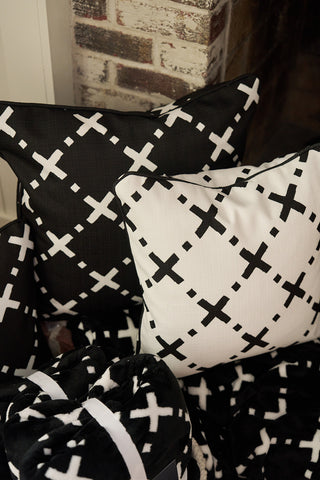 Two cozy square pillows with black and white X-patterns rest against a brick wall, with The Paige Collection: Exes in Black Blanket partially visible at the bottom for added warmth and style.