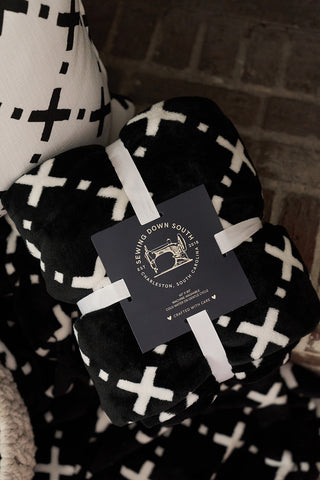The Paige Collection: Exes in Black Blanket is a folded, extra plush black blanket with white X patterns, tied with a white ribbon. A Sewing Down South tag with a sewing machine logo is attached, and it rests by a matching patterned pillow.