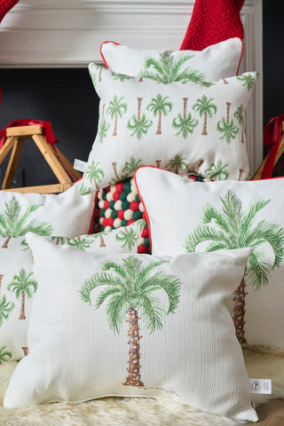 The Holiday Palmetto Lumbar Pillow, featuring green palm tree designs on white, is styled in a cozy scene with a furry rug, woven basket with red ribbons, and a fireplace adorned with red stockings.