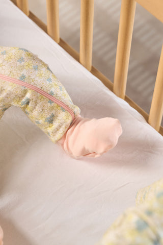 A close-up of a baby’s legs in the Precious Petals Dream & Play Zipper Romper, with one foot in a soft pink sock, resting on a white crib sheet beside light wooden crib rails.