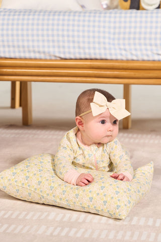 A baby wearing a large cream bow headband and patterned onesie lays on a Precious Petals Children’s Sleeping Pillow Cover—machine washable and matching her outfit—on a beige rug, with a wooden bench and gingham cushion in the background.