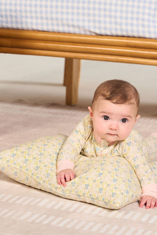 A baby lies on a patterned, machine washable Precious Petals Children’s Sleeping Pillow Cover on a beige rug, looking up. Behind the baby is a wooden bench with blue and white checkered cushions and pillows featuring double-sided prints of charming bee designs. A woven basket sits to the side.