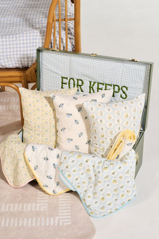 An open suitcase labeled FOR KEEPS displays the Buggin’ Out Children’s Sleeping Pillow Cover and patterned blankets with bees, hearts, and florals beside a rattan chair and blue gingham bedspread.