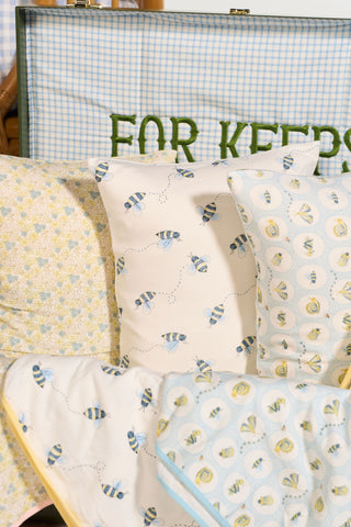 Several patterned pillows, including the Buzzin’ Around Children’s Sleeping Pillow Cover and cozy floral pillow covers, are arranged on a bed. Behind them is a blue checkered case with green text partially reading FOR KEEPS.