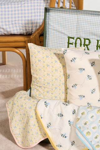 A close-up of the Precious Petals Children’s Blanket, featuring bee and floral patterns on bedding, alongside a wicker chair topped with a blue-checked decorative throw pillow, and part of an open suitcase showcasing green “FOR K” embroidery.