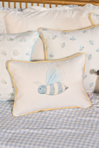 The Bee You Children’s Lumbar Pillow features a blue and yellow bee design and double-sided prints, shown here on a bed with blue gingham sheets alongside other bee-themed pillows for a playful, coordinated look.