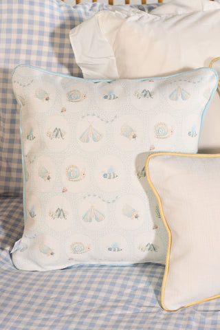 The Buggin’ Out Children’s Pillow, square with light blue piping and a bug print of bees, butterflies, and snails, sits on blue gingham bedding. A white pillow with yellow piping is partially visible in front.