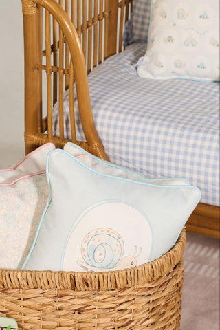 A wicker basket with embroidered pillows sits next to a rattan crib with blue and white checkered bedding and a Buggin’ Out Children’s Pillow in a cozy nursery setting.