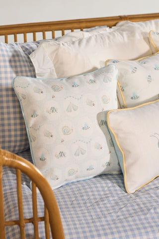 Square throw pillows with light blue and white seashell and starfish patterns rest on a blue gingham bedspread, alongside the Buggin’ Out Children’s Pillow and other white pillows with yellow piping on a wooden bedframe.
