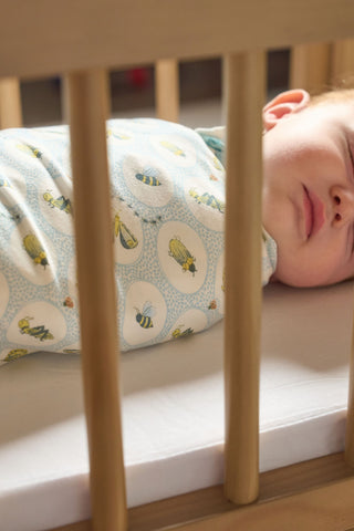 A baby sleeps on its back in a crib, wrapped in the Buggin’ Out Children’s Blanket, which features white and blue colors with bee patterns. Crib bars are visible in the foreground.
