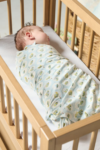 A newborn sleeps on their side in a wooden crib with light bedding, swaddled in the Buggin’ Out Children’s Blanket featuring a fun bug print.