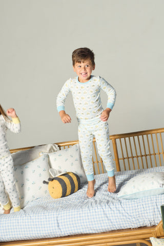 A young child in Buggin’ Out Dream & Play Two Piece Set cotton modal pajamas jumps on a bed with blue gingham sheets and a plush bee pillow; another child stands partly out of frame.