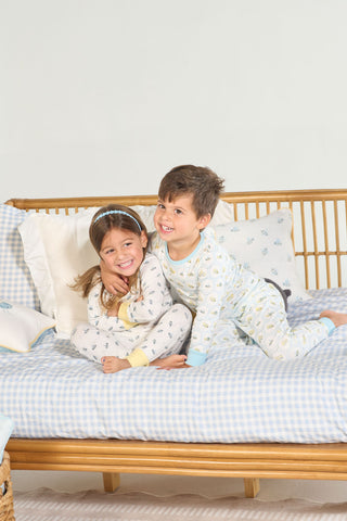 Two young children in Buzzin’ Around Dream & Play Two Piece Set smile and pose playfully on a rattan daybed with blue-and-white checkered bedding.