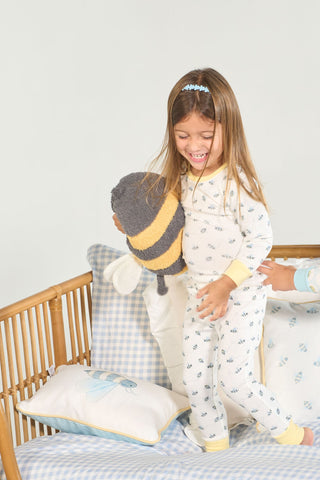 Young child in the Buzzin’ Around Dream & Play Two Piece Set stands on a bed holding a stuffed bee, smiling. Blue and white checked bedding with a bee pillow adds to the cozy cotton modal pajama scene.