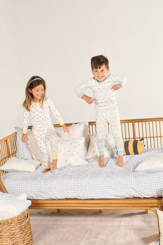 Two children wearing Buggin’ Out Dream & Play Two Piece Set pajamas laugh and jump on a wicker daybed with blue-checkered bedding and pillows against a plain, light-colored background.