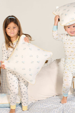 Two young children in matching pajamas have a pillow fight, smiling and holding pillows. Buzzin’ Around Children’s Pillow is featured among the light bedding and playful children’s decor pillows.