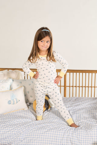 A young girl in a Buzzin’ Around Dream & Play Two Piece Set stands confidently on a bed with blue plaid bedding, hands on hips and a playful expression. Bee-themed pillows decorate the bed behind her.