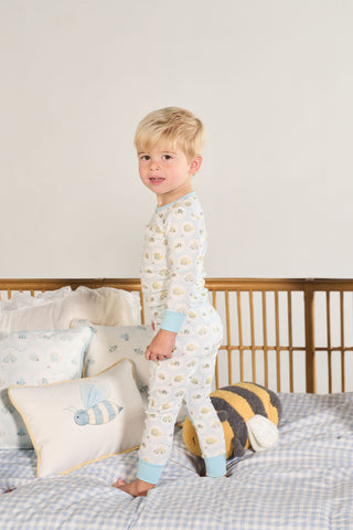 A young child in the Buggin’ Out Dream & Play Two Piece Set smiles on a bed with bee-themed pillows and a plush bee, with a wooden crib and light-colored walls in the background.