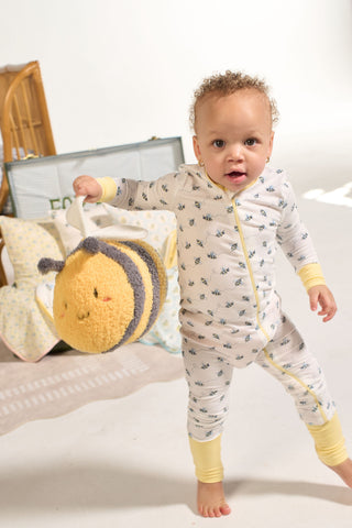 A toddler in a Buzzin’ Around Dream & Play Zipper Romper stands barefoot holding a plush bee bag, with pillows and a wicker basket in the soft, bright background.