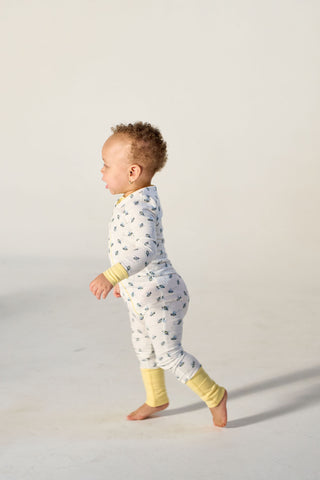 A toddler in the Buzzin’ Around Dream & Play Zipper Romper with yellow cuffs walks barefoot on a light floor, facing left, against a neutral background.