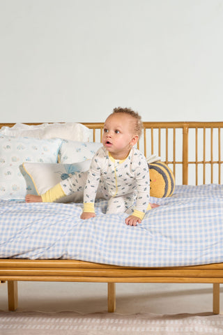 A baby in a Buzzin’ Around Dream & Play Zipper Romper sits on a bed with blue checkered sheets, surrounded by pillows—including a round striped one—and a wooden headboard in the background.