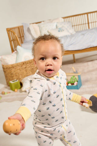 A toddler in the Buzzin’ Around Dream & Play Zipper Romper holds a toy egg, standing in a playroom with a wicker crib and toys in the background.