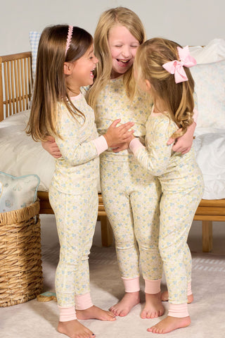 Three young girls in matching Precious Petals Dream & Play Two Piece Set pajamas stand on a rug by a bed, hugging and smiling happily. Nearby, you can spot a made in USA pillow that’s both stylish and comfortable, along with a wicker basket and a toy shelf filled with books and toy vegetables.
