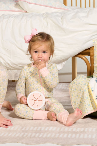 A toddler wearing the Precious Petals Dream & Play Zipper Romper with a pink bow sits on a rug, holding a round object. Behind her is a bed with white bedding; she looks at the camera, hand near her mouth.