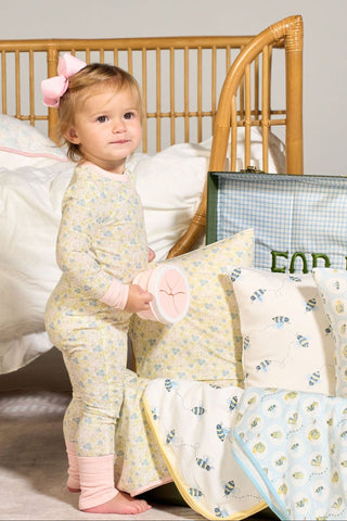 A toddler with a pink bow stands by a bed, wearing the Precious Petals Dream & Play Zipper Romper, holding a round toy among blue bee pillows, blankets, a woven chair, and a green-striped bag.