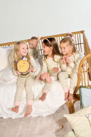 Four young children in matching Precious Petals Dream & Play Two Piece Sets sit and smile on a bed with white bedding. One child holds a plush bee toy while cozy pillows, adorned with playful double-sided prints, rest on the floor nearby. The bed’s wicker frame adds to the cheerful and inviting scene. All textiles shown are machine washable for easy care, making this playful moment both fun and practical.