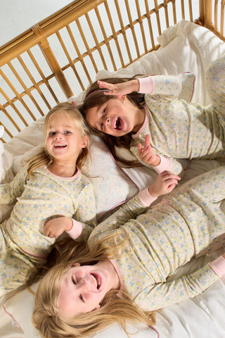 Three young children wearing matching Precious Petals Dream & Play Two Piece Sets laugh and play together on a bed with white bedding, next to a wooden crib headboard and a decorative throw pillow.