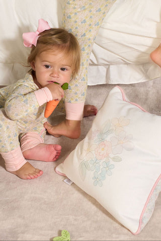 A toddler with a pink bow holds a toy carrot while sitting on a soft surface next to the Precious Petals Children’s Pillow, with an adult’s legs in matching pajamas nearby.