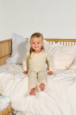 A young girl with long blonde hair, wearing the Precious Petals Dream & Play Two Piece Set, sits smiling on a white bed with a wicker frame. Behind her are several pillows, including a blue gingham decorative pillow. The room has a soft, cozy atmosphere.