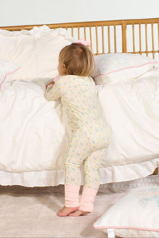 A toddler in a Precious Petals Dream & Play Zipper Romper and pink leg warmers stands at the edge of a bed, facing the bedding with hands resting on the blanket.