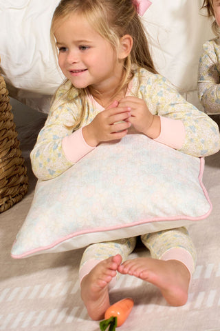 A young girl in patterned pajamas sits on the floor, smiling and hugging the Precious Petals Children’s Pillow with double-sided prints. Her feet are bare, with a toy carrot near her toes. Another child is partially visible beside her.