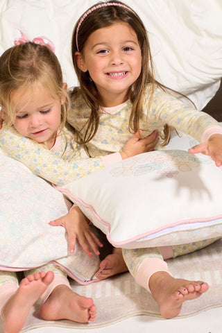 Two young girls in pajamas sit on the floor in front of a bed, smiling as they hold the Precious Petals Children’s Pillow. The scene feels cozy with white bedding and soft lighting, while the playful double-sided prints on the pillow add a charming touch.