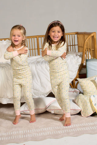 Two young children, barefoot and smiling, stand next to a bed wearing the Precious Petals Dream & Play Two Piece Set. The cozy scene includes a rattan bed with white bedding and a decorative pillow, with extra pillows and blankets scattered on the floor.