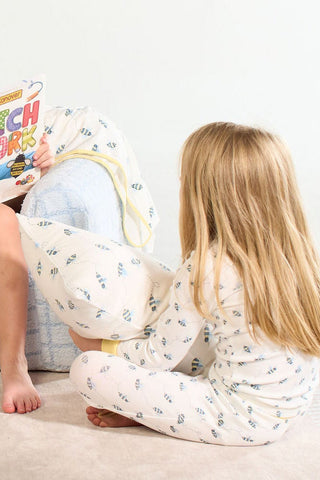 A child in fish-patterned pajamas sits on the floor, holding the Buzzin’ Around Children’s Sleeping Pillow Cover, facing another child reading a book in a cozy, soft atmosphere.