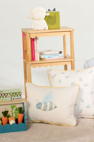 A small wooden shelf holds books, a sheep lamp, and a green radio. On the floor are decorative pillows featuring bee designs—including the Bee You Children’s Lumbar Pillow, which is double-sided and made in the USA—and a wooden toy box filled with play vegetables.