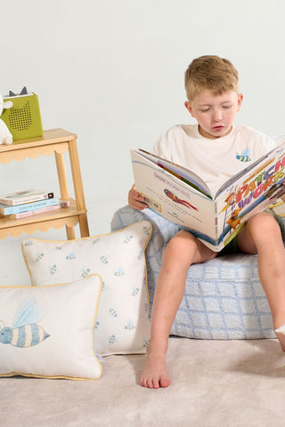 Young boy sits on a light blue cushion reading a book. A small wooden shelf, books, green basket, and the Buzzin’ Around Children’s Pillow add cozy charm to the bright scene.