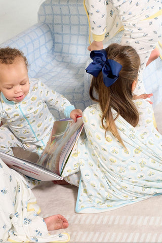 Two kids in pajamas sit on a soft rug, one draped in the Buggin’ Out Children’s Blanket. One holds an open book, while the other wears a big blue bow. A third child stands partially out of frame.