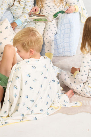 Four children in bee-print pajamas read together under the Buzzin’ Around Children’s Blanket, lounging on a soft rug and blue chair in a bright, cozy room.